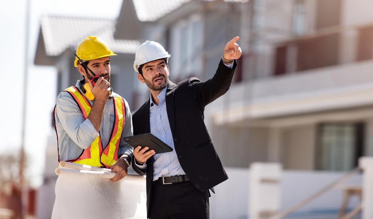 Two tradesmen in hard hats, one in a safety vest with a radio, reviewing plans on a building site
