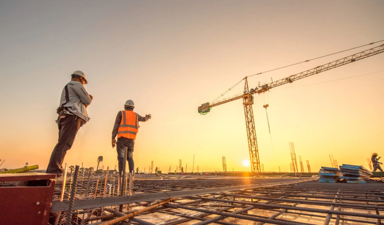 Two engineers in hard hats on a construction site at sunset with a tower crane in the background