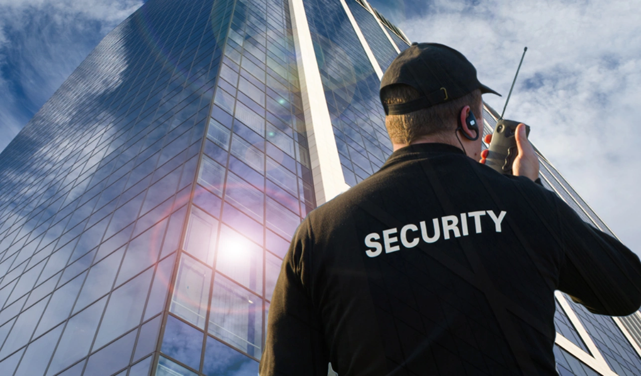 Security officer in a black SECURITY jacket speaking into a two-way radio outside a glass office building
